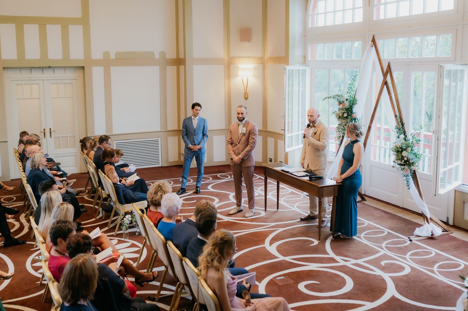 Intimate indoor wedding ceremony with guests seated in rows, officiant and couple at wooden arch with floral decor.