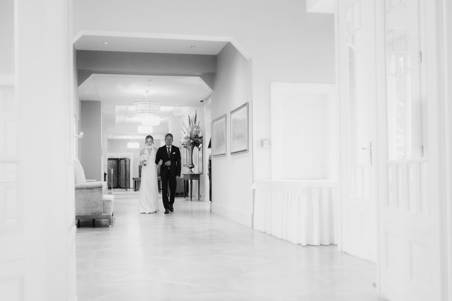 Black and white photo of couple walking through bright hallway at wedding venue.