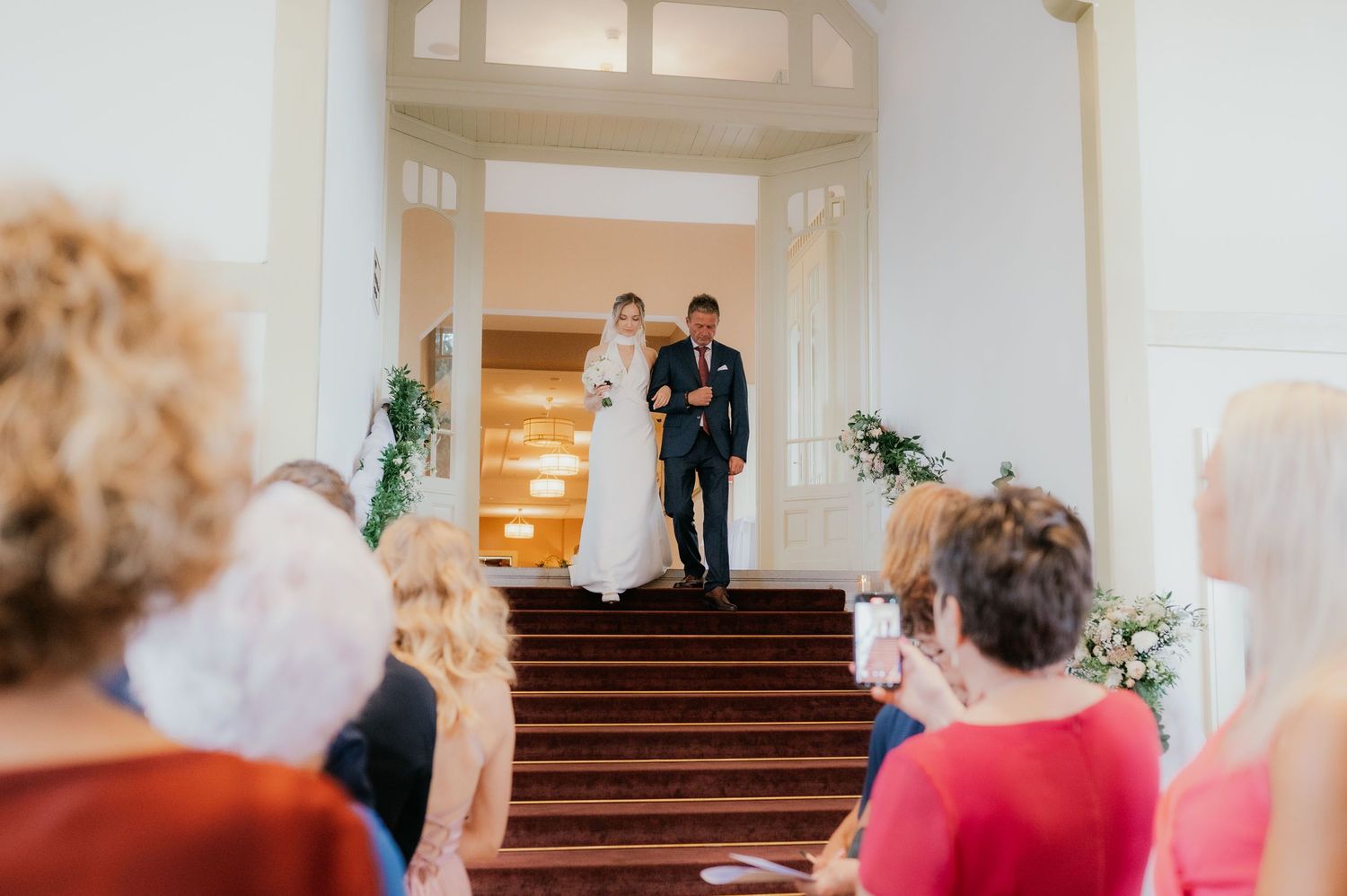 Bride and groom standing at top of stairs during wedding ceremony with guests seated in foreground.