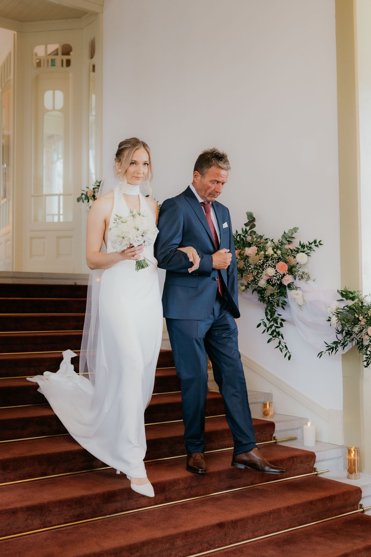 Bride in white gown walks down red-carpeted stairs with man in navy suit at elegant wedding venue.
