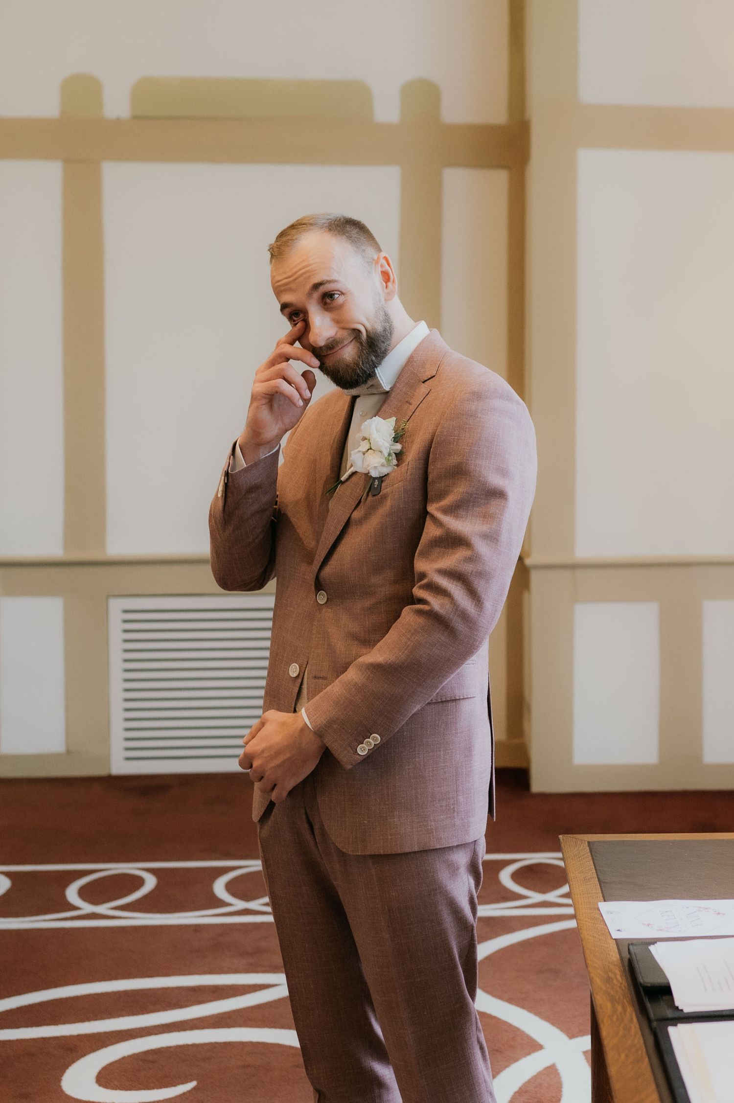 Emotional groom in brown suit wiping tears with boutonniere on lapel during wedding ceremony indoors.