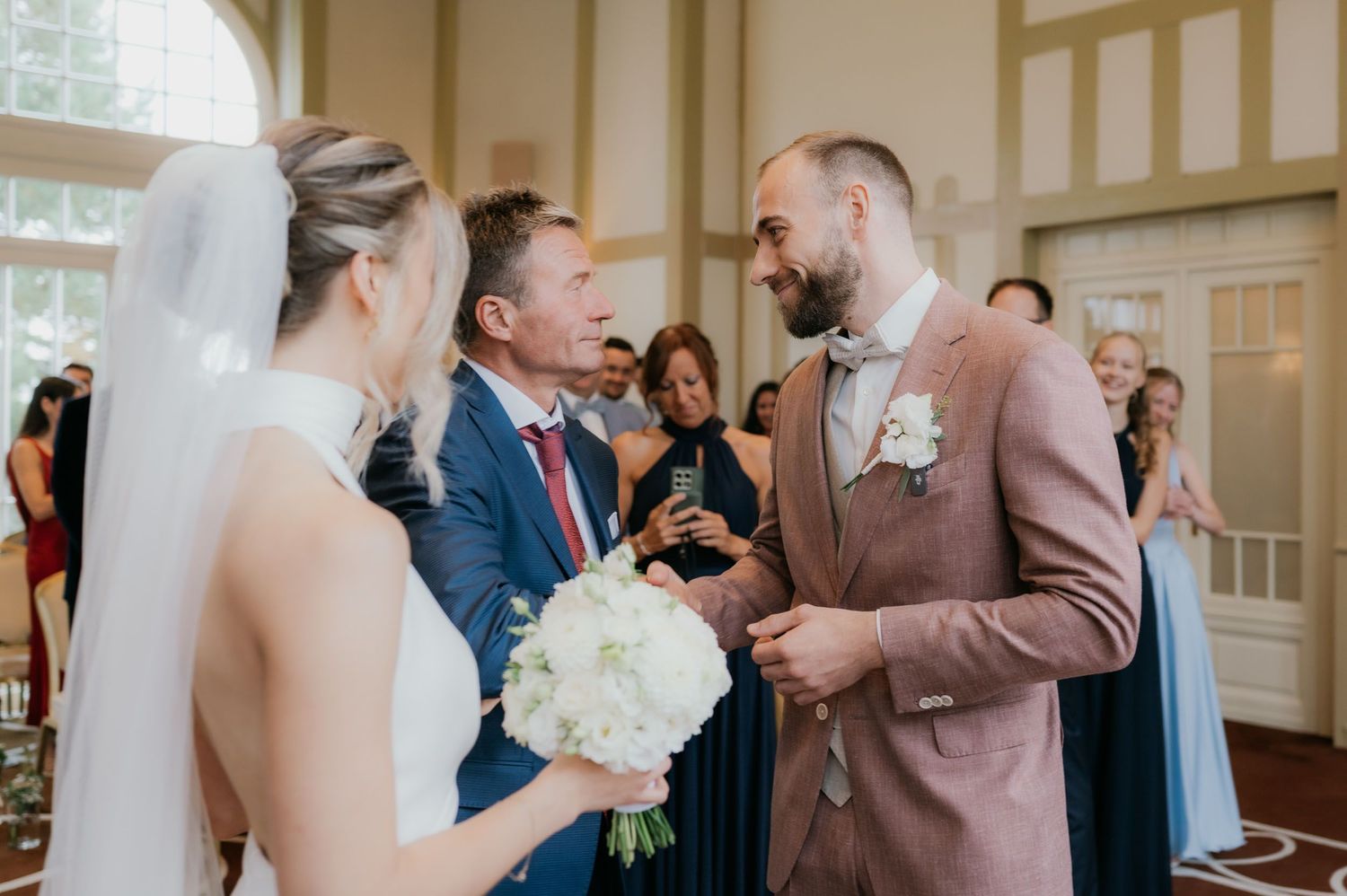 Bride and groom exchanging vows at elegant wedding ceremony, groom in pink suit holding bride's hands.