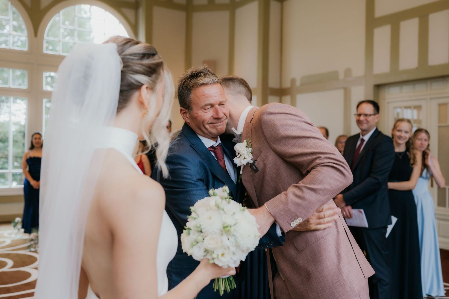 Groom in pink suit hugs man at wedding ceremony while bride in white gown holds bouquet of white flowers.
