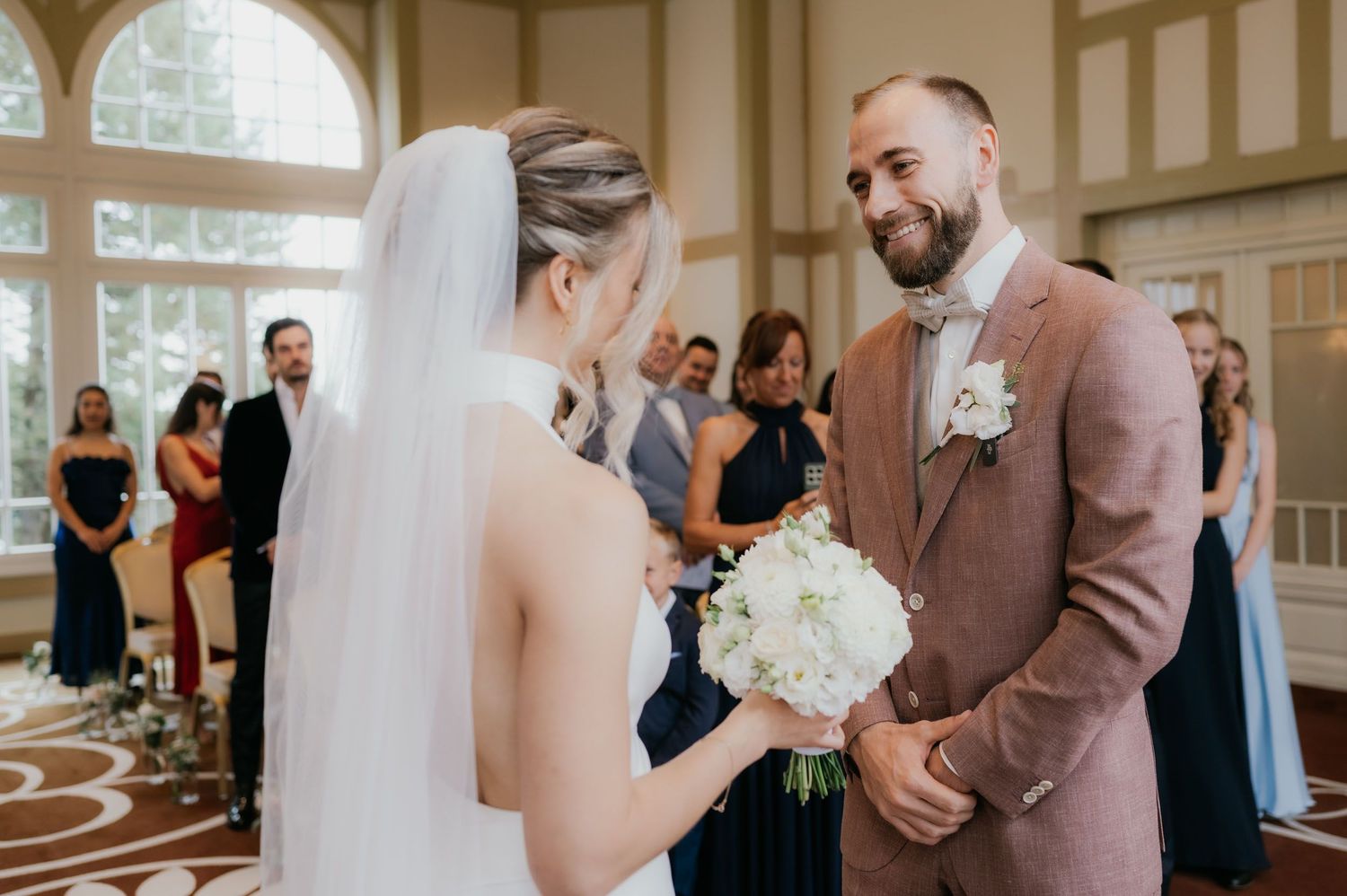 Groom in pink suit smiling at bride holding white bouquet during indoor wedding ceremony.