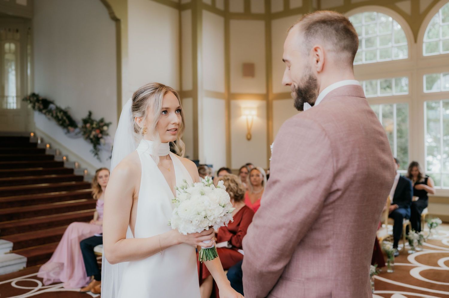Bride in white gown smiling at groom during elegant indoor wedding ceremony with guests seated.