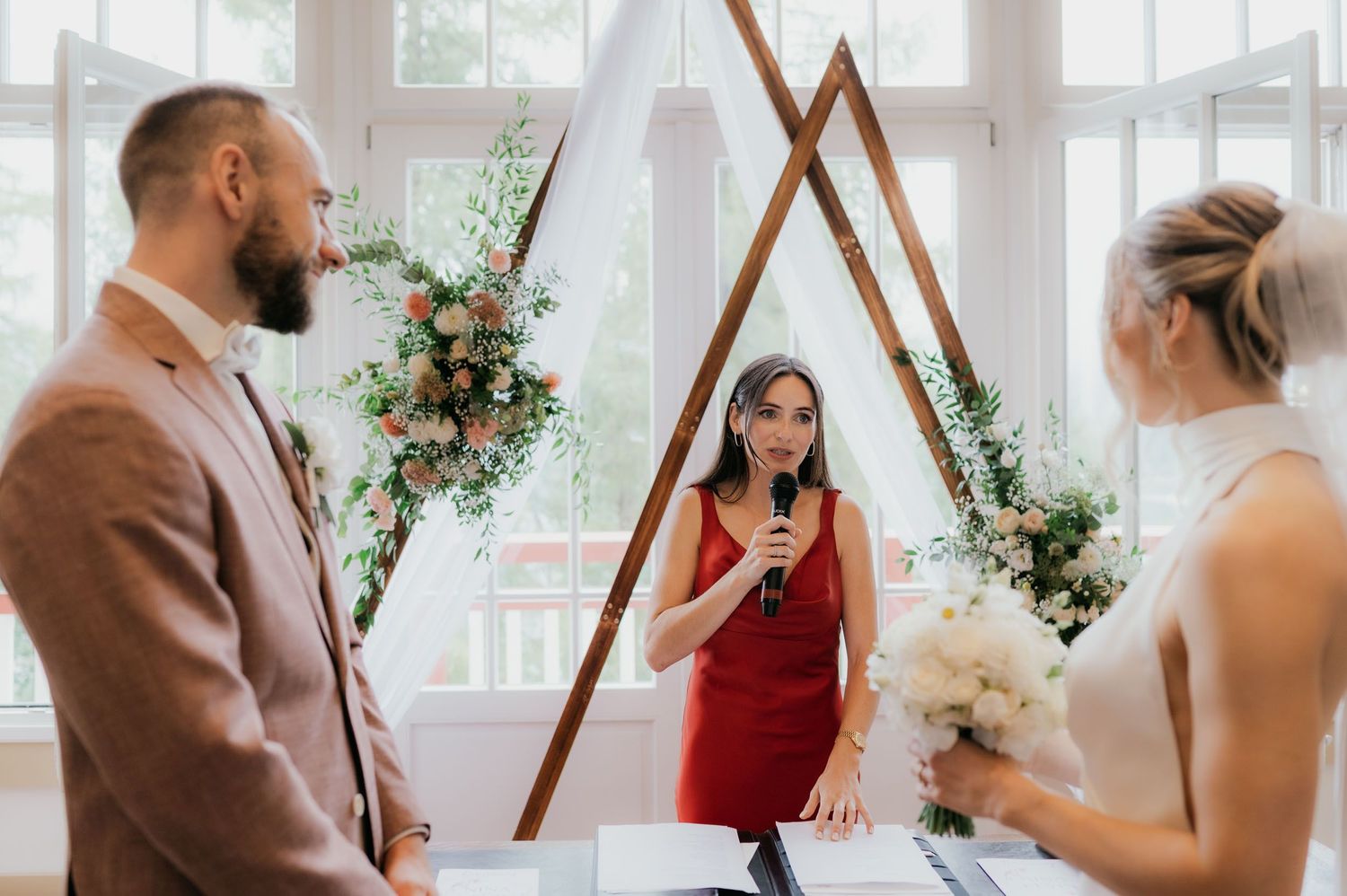 Wedding ceremony with officiant in red dress speaking between couple at triangular wooden arch with floral arrangements.