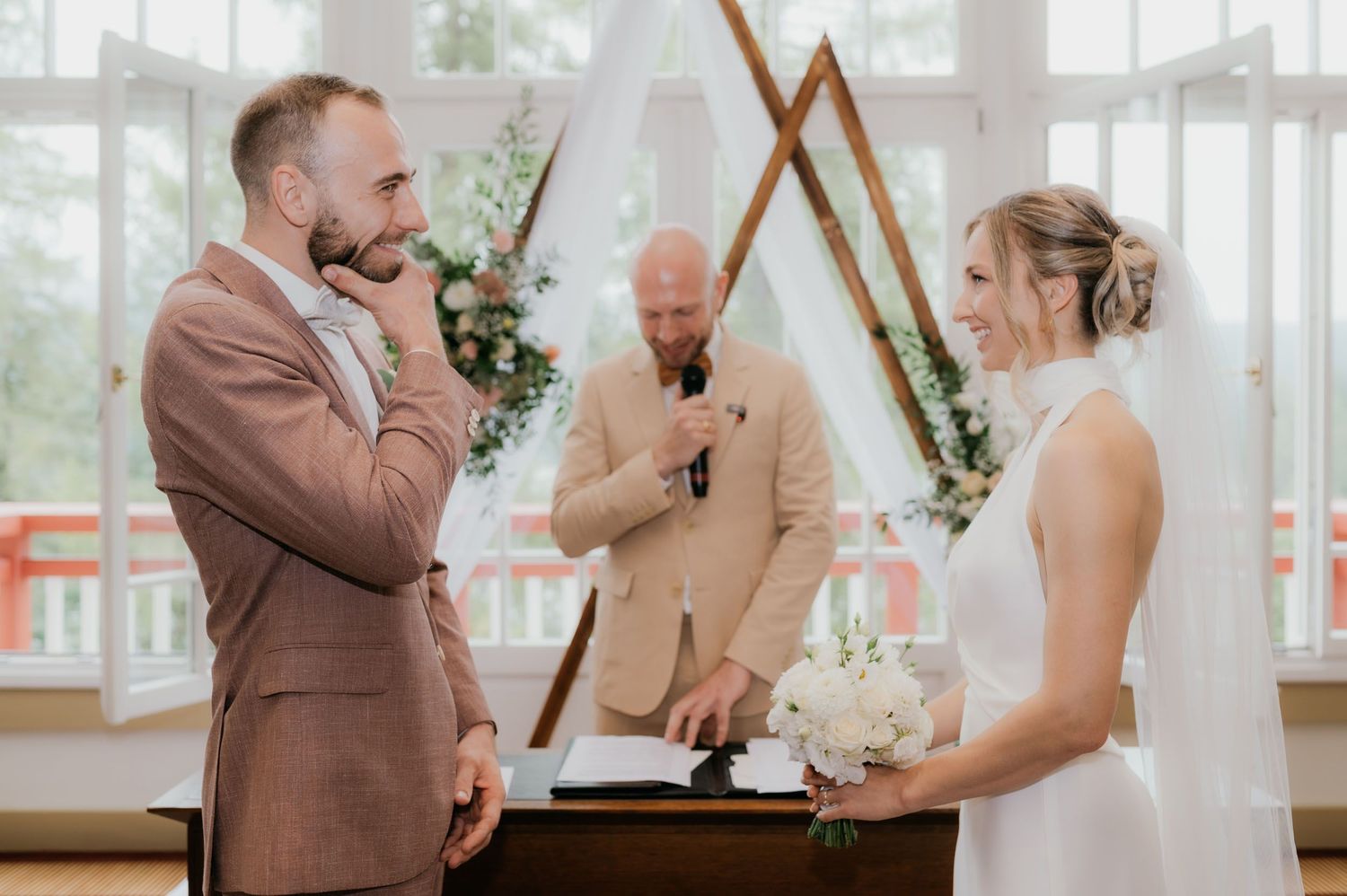 Emotional groom listening to wedding vows at an indoor ceremony with a geometric arch backdrop and officiant.