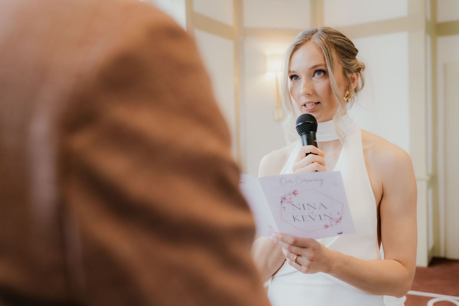 Bride in white dress giving speech at wedding ceremony, holding microphone and floral program card.