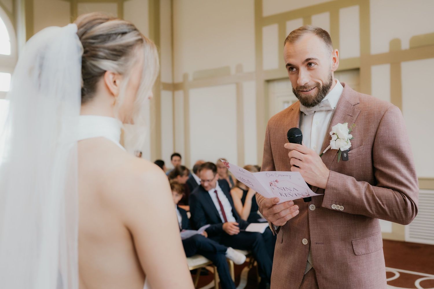 Groom reads wedding vows to bride during indoor ceremony, wearing pink suit with boutonniere.