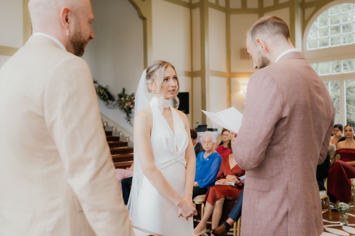 Bride and groom exchange vows at elegant indoor wedding ceremony with guests seated in background.