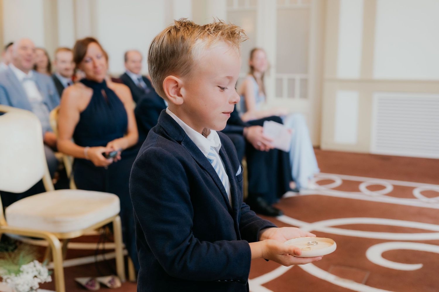 Young ring bearer in navy suit carefully holding wedding rings during an elegant indoor ceremony.