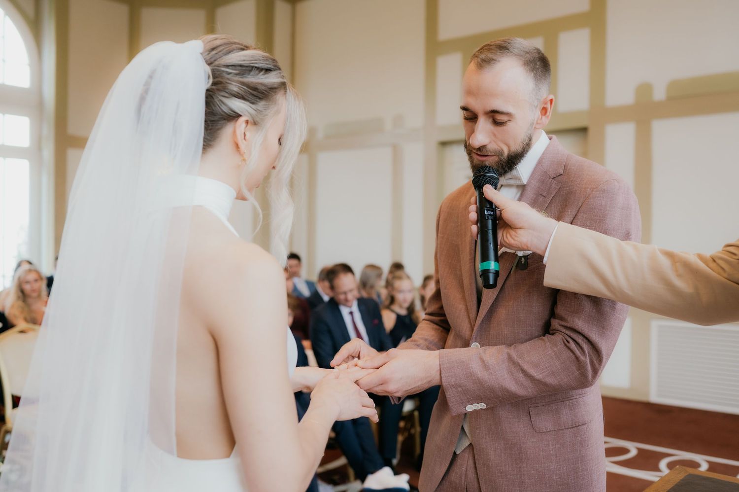 Groom placing wedding ring on brides finger during ceremony while officiant holds microphone nearby.