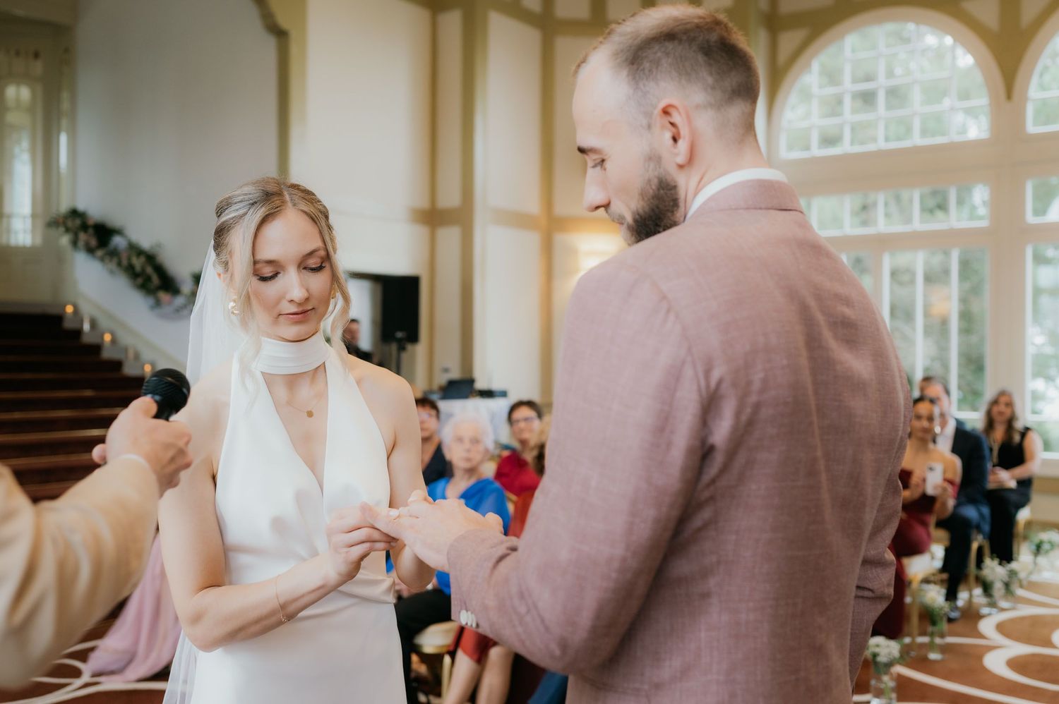 Bride and groom exchanging rings during wedding ceremony in elegant venue with guests seated in background.