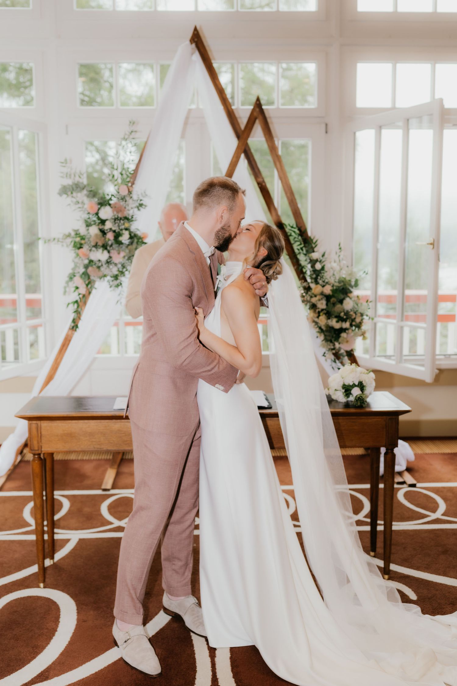Bride and groom share first kiss at elegant indoor wedding ceremony with geometric arch and floral arrangements.