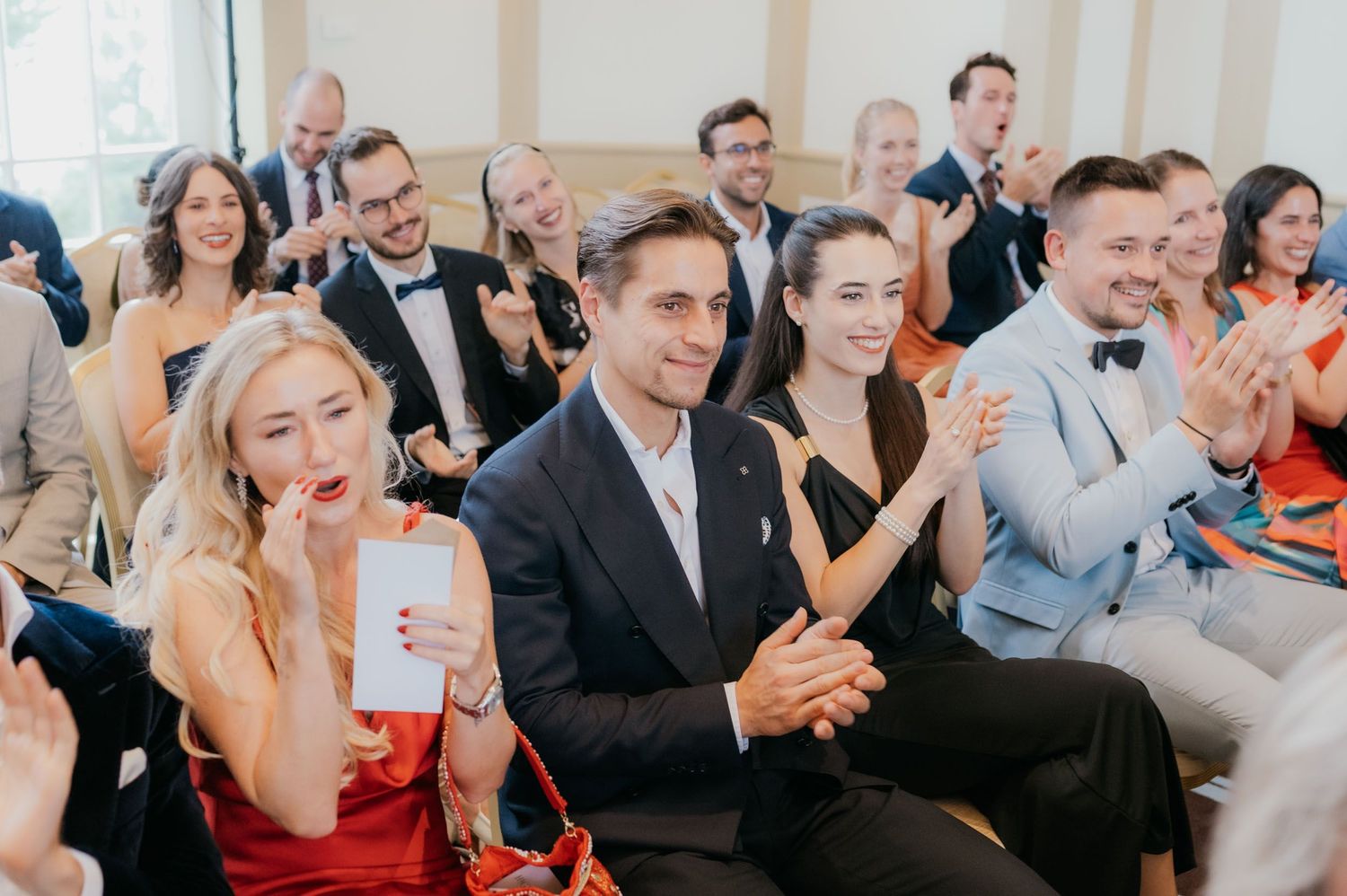 Wedding guests in formal attire clapping and smiling during a ceremony in an elegant venue.