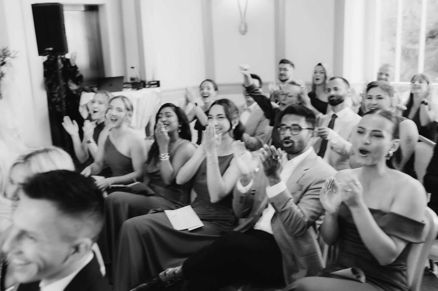 Black and white photo of excited wedding guests cheering and applauding during a reception.