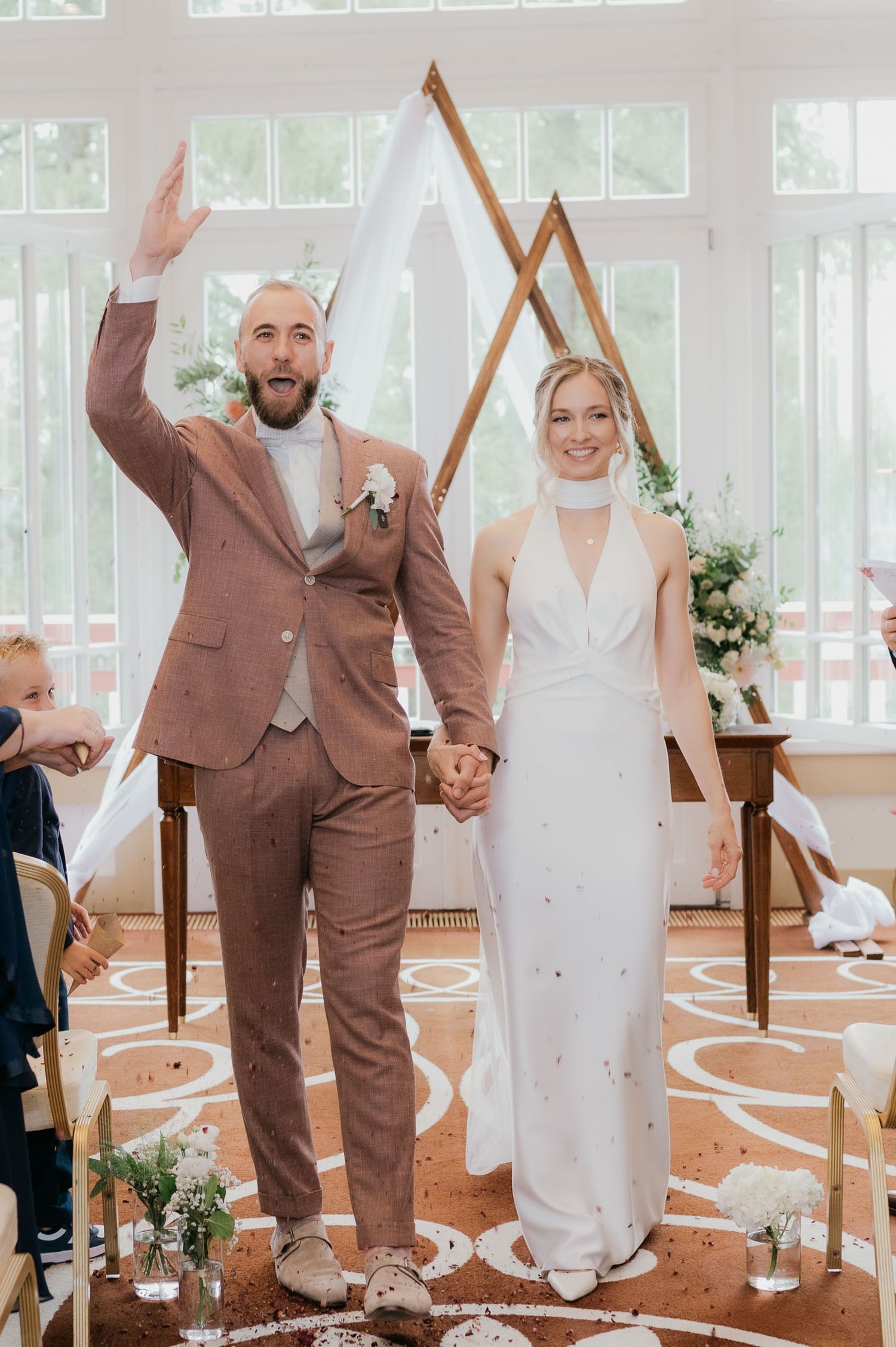 Joyful newlyweds hold hands at wedding altar, groom in brown suit raises arm, bride in white halter gown, geometric backdrop.