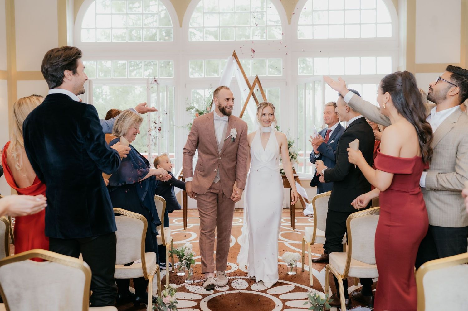 Bride and groom walk down the aisle as guests celebrate with confetti at an elegant indoor wedding ceremony.