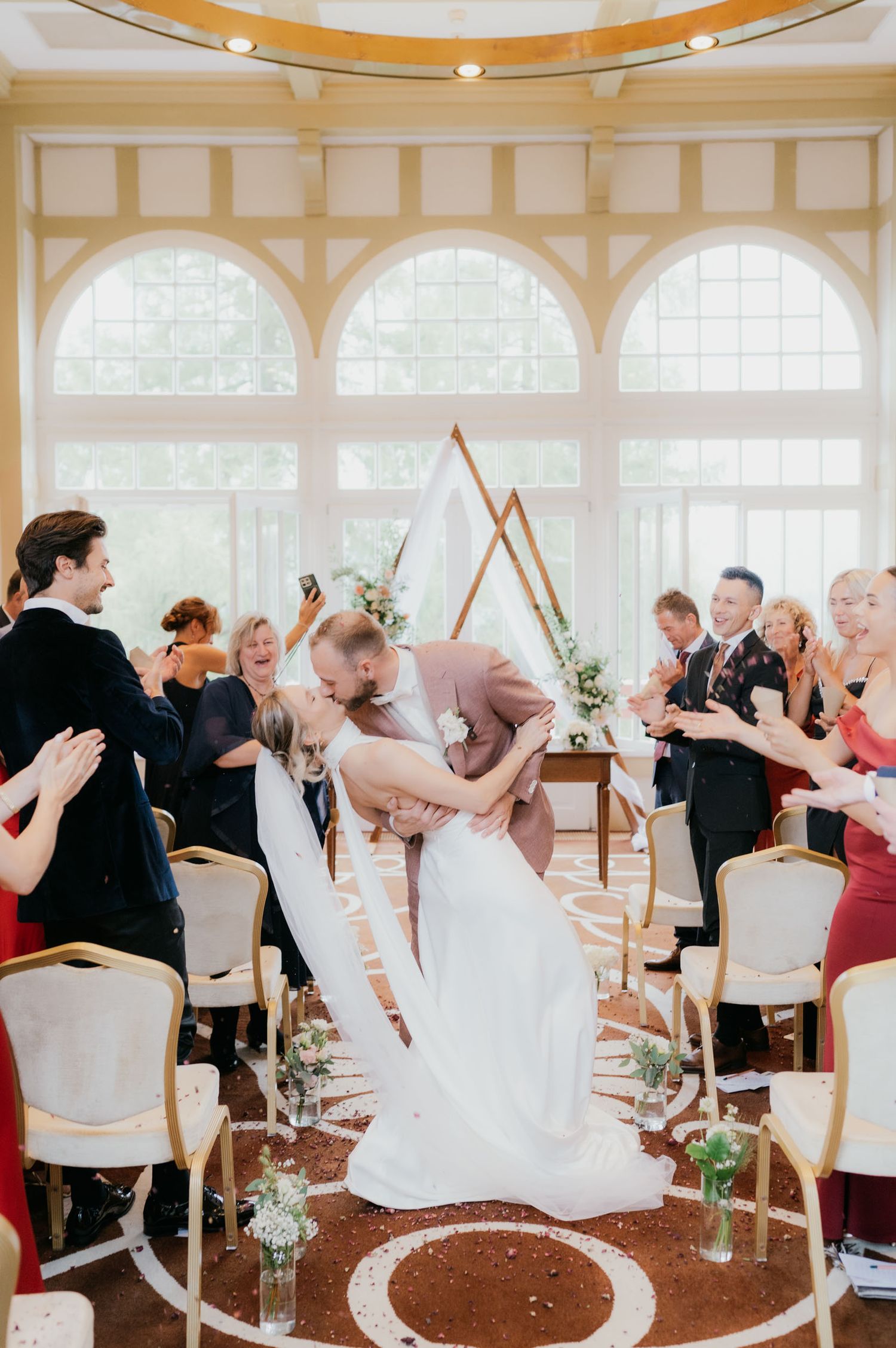Groom dips bride for a kiss during wedding ceremony as guests clap and toss confetti in elegant venue.