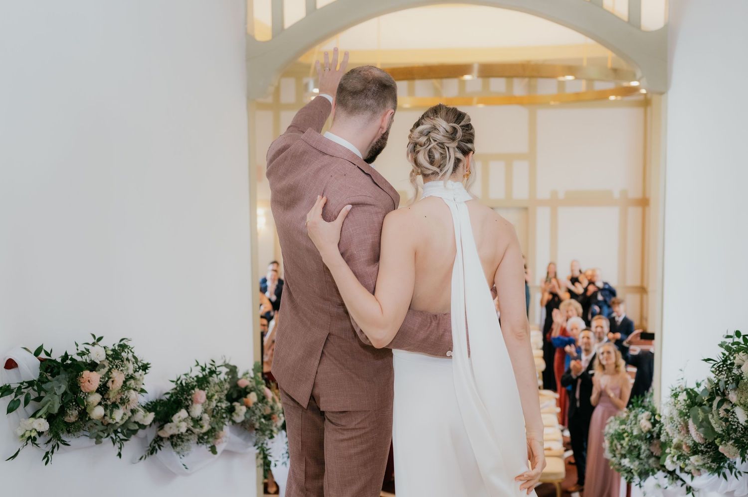 Bride and groom standing at wedding altar, viewed from behind, waving to guests in elegant venue with floral arrangements.