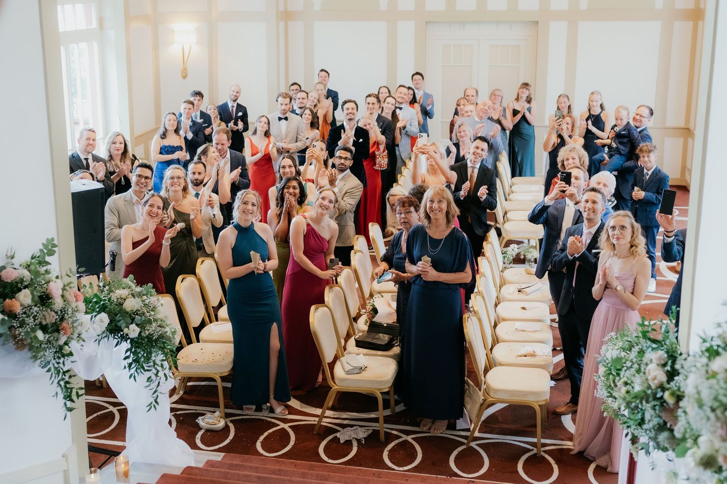 Wedding guests clapping and celebrating in an elegant ballroom decorated with floral arrangements and gold chairs.