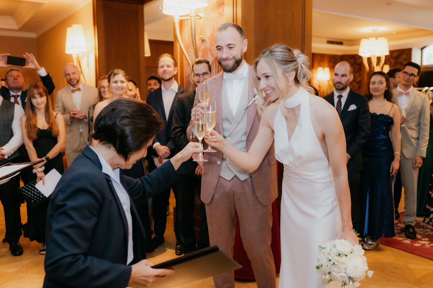 Bride and groom toasting champagne glasses at wedding reception surrounded by smiling guests in elegant venue.