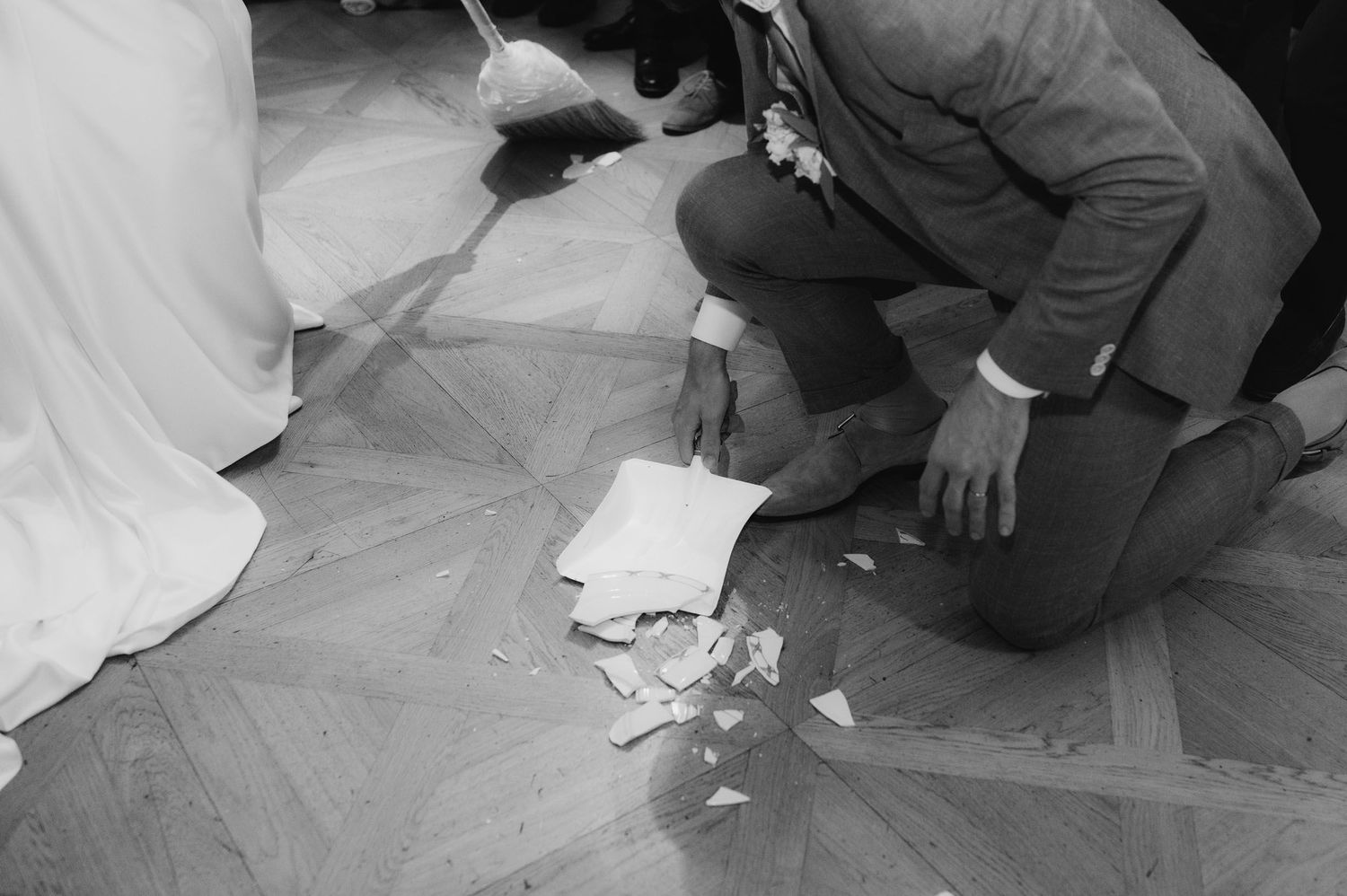 Black and white photo of groom kneeling to pick up broken glass on herringbone wood floor at wedding ceremony.