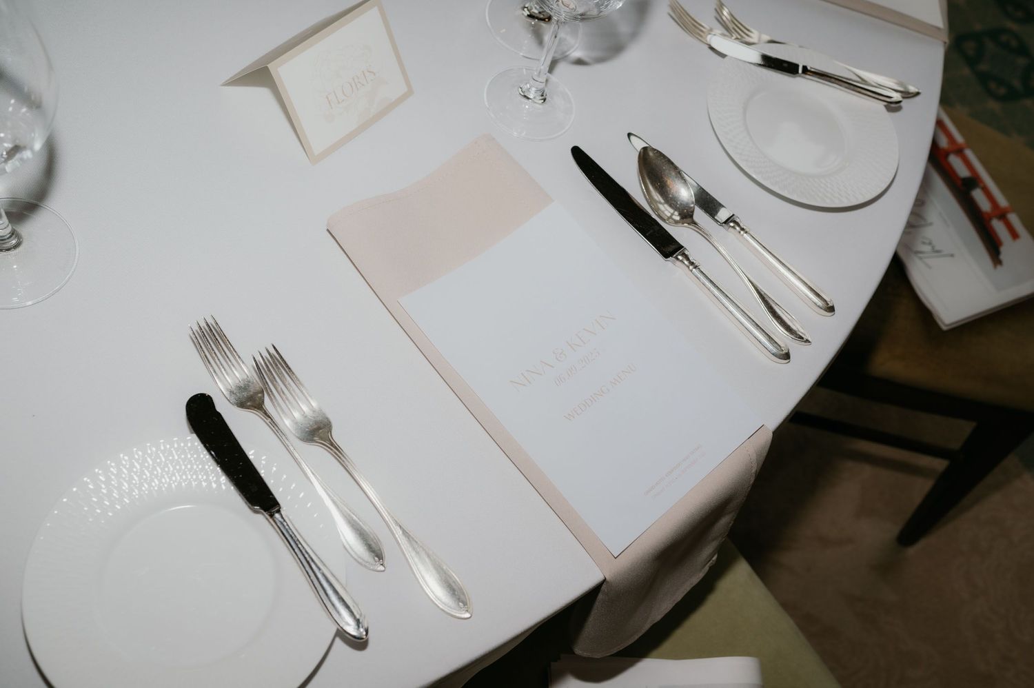 Elegant wedding reception table setting with white plates, silverware, menus, and glassware on a round white tablecloth.