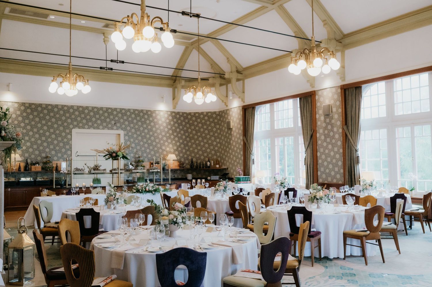 Elegant wedding reception hall with round tables, white linens, floral centerpieces, and gold chandeliers in a bright venue.