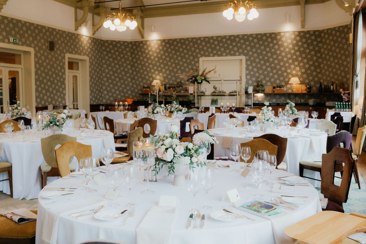 Elegant wedding reception hall with round tables, white linens, floral centerpieces, and classic wooden chairs in a decorated venue.