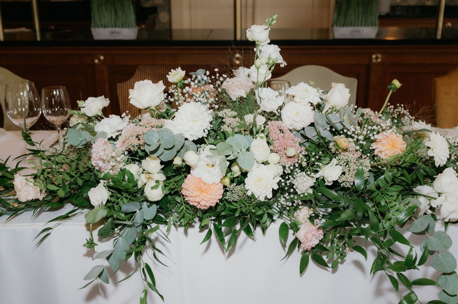 Elegant wedding table centerpiece with white and blush roses, eucalyptus greenery, and baby's breath on a white tablecloth.