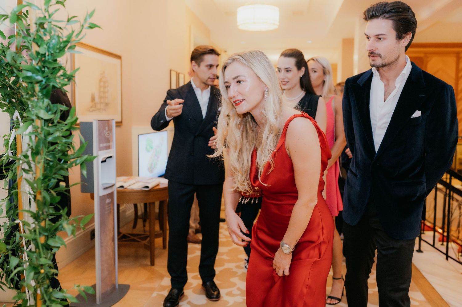 Elegantly dressed guests in formal attire gather indoors at a sophisticated event, with a woman in red standing prominently.