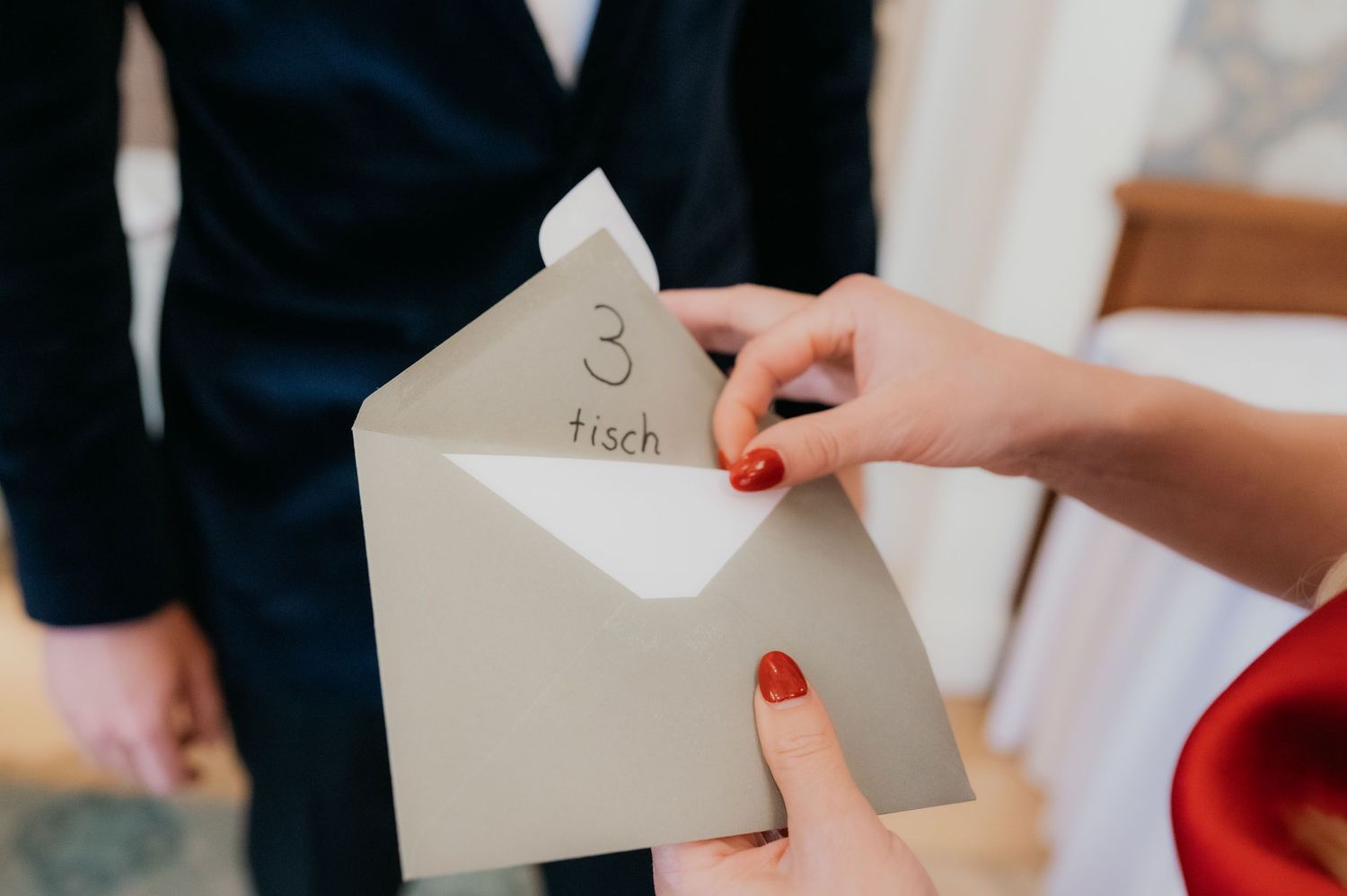 Hand with red nails pulling a table number card from a white envelope at a wedding reception.