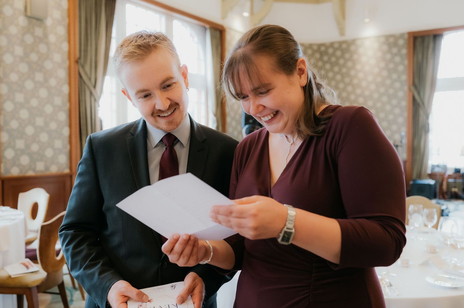 A young man in a suit and woman in burgundy dress smile while reviewing a document together in an elegant restaurant.