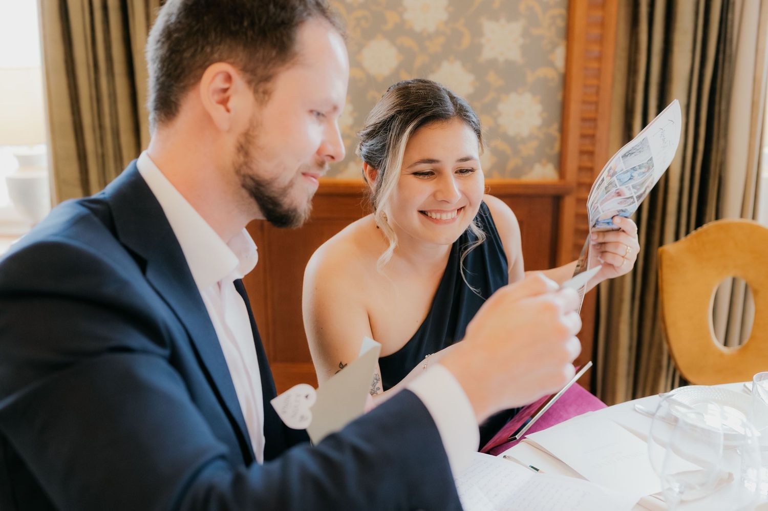 Couple smiling and reading a card together at a wedding reception table in an elegant dining room.