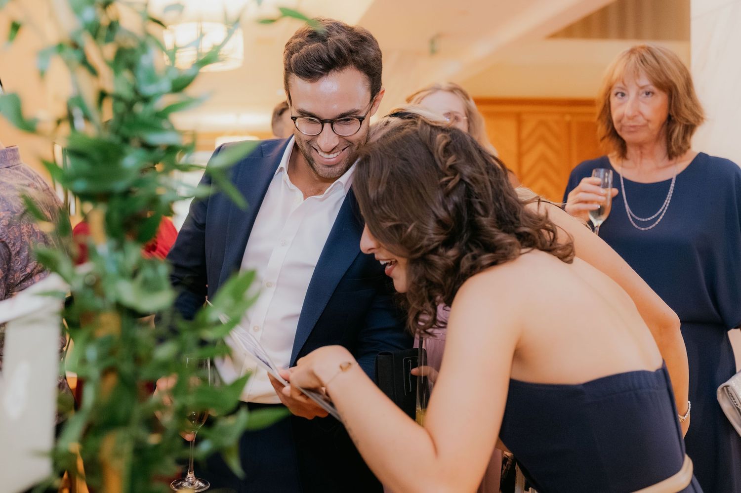 A smiling man in a navy suit and glasses looks at a woman writing on a card at an elegant wedding reception.