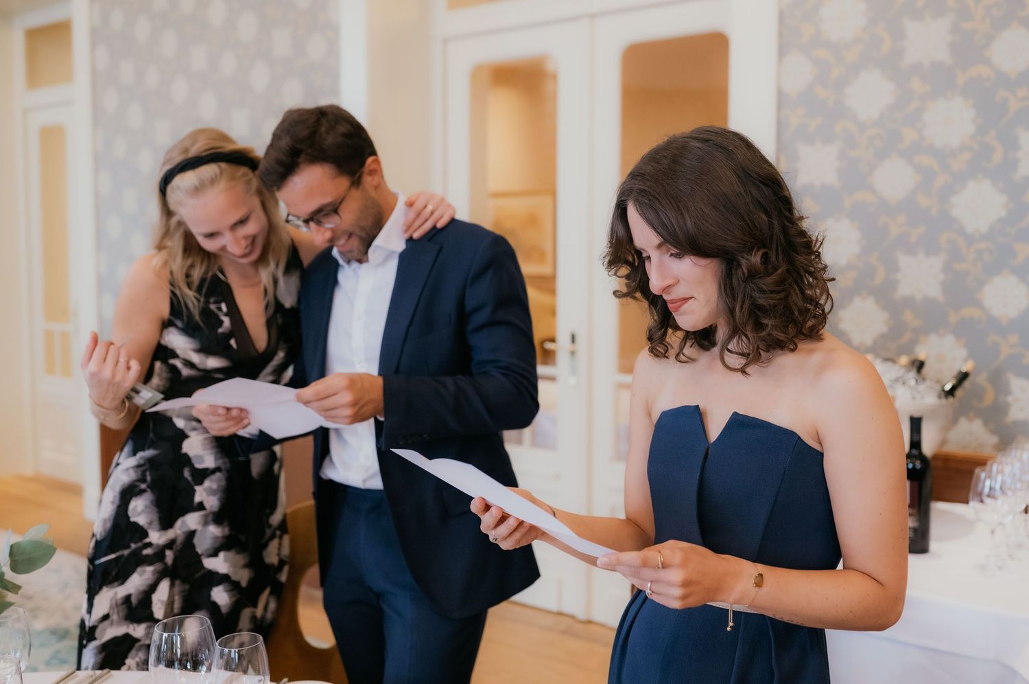 Three elegantly dressed guests reading papers at a formal wedding reception in a decorated venue.