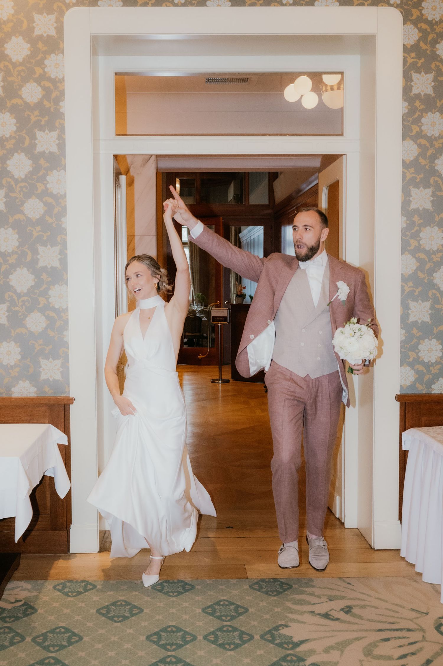 Bride and groom make grand entrance holding hands raised in elegant venue with patterned wallpaper and hardwood floors.