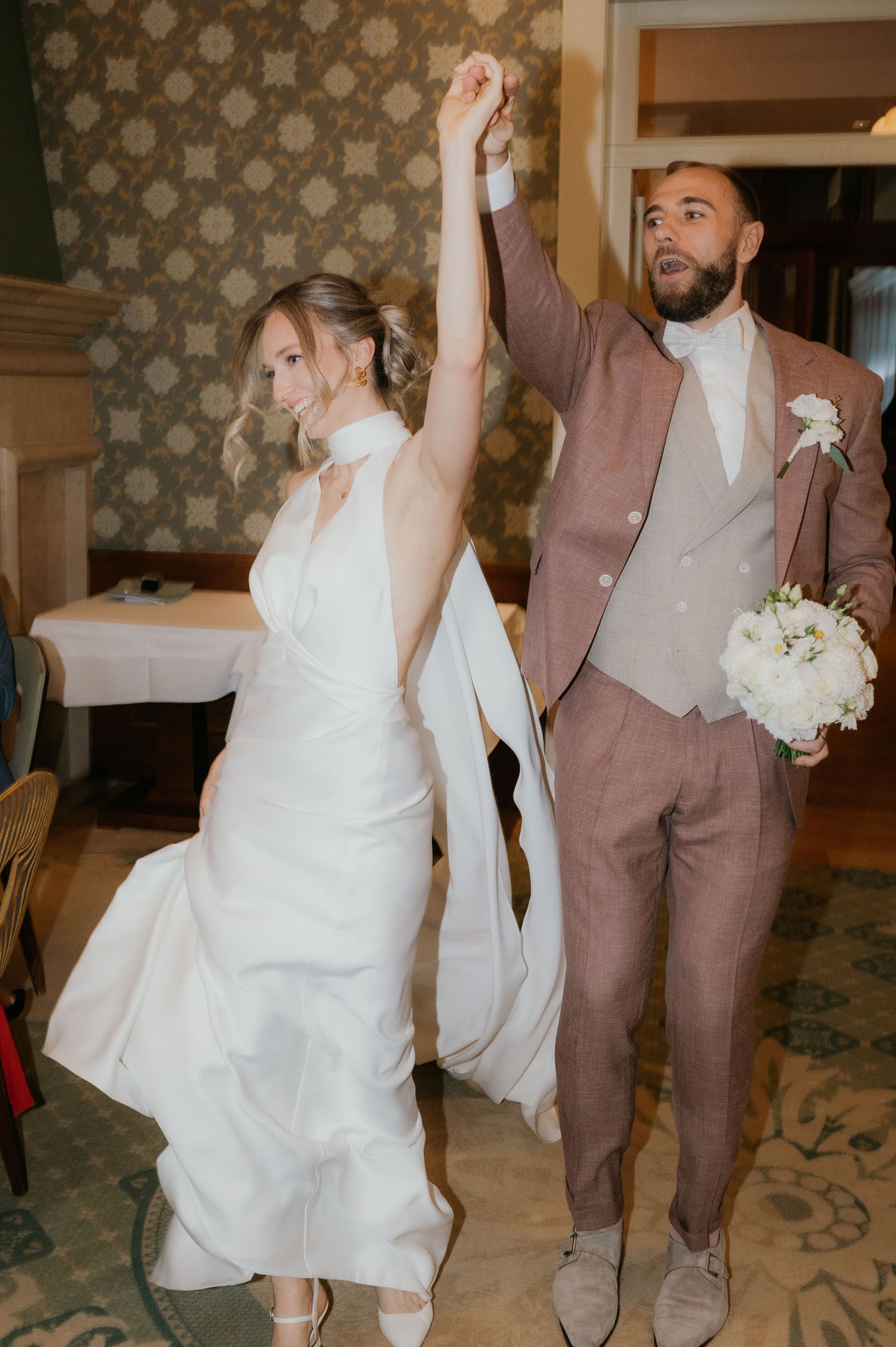 Joyful newlyweds celebrate their wedding entrance, bride in white gown and groom in mauve suit raising hands triumphantly.