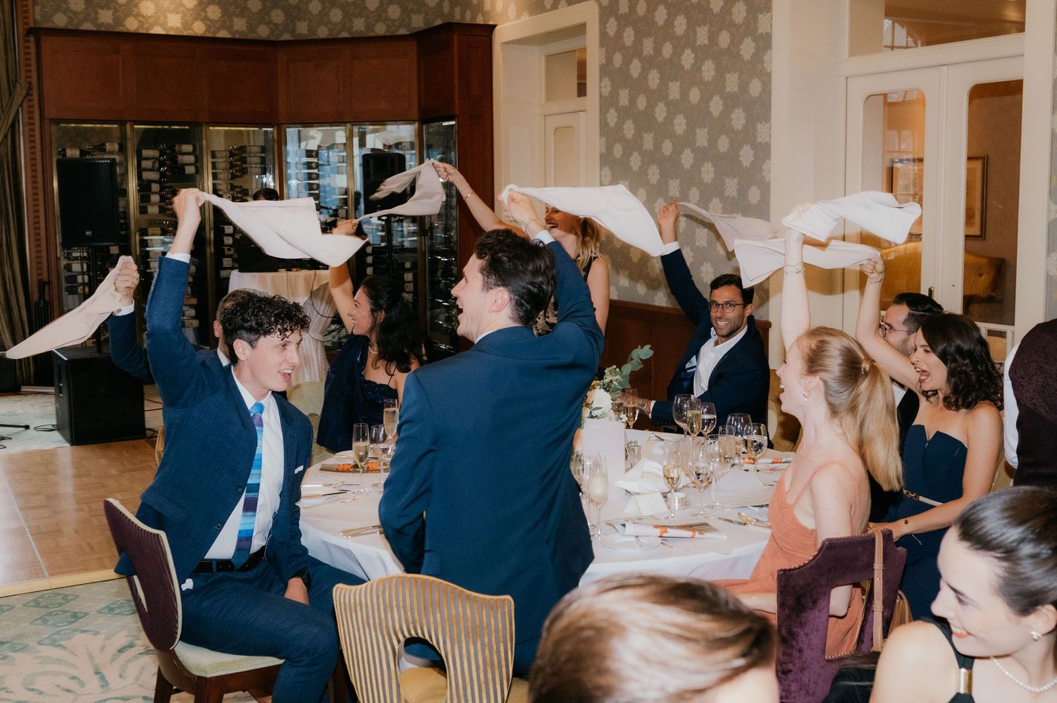 Guests wave white napkins in celebration at an elegant wedding reception dinner in a warmly lit dining room.