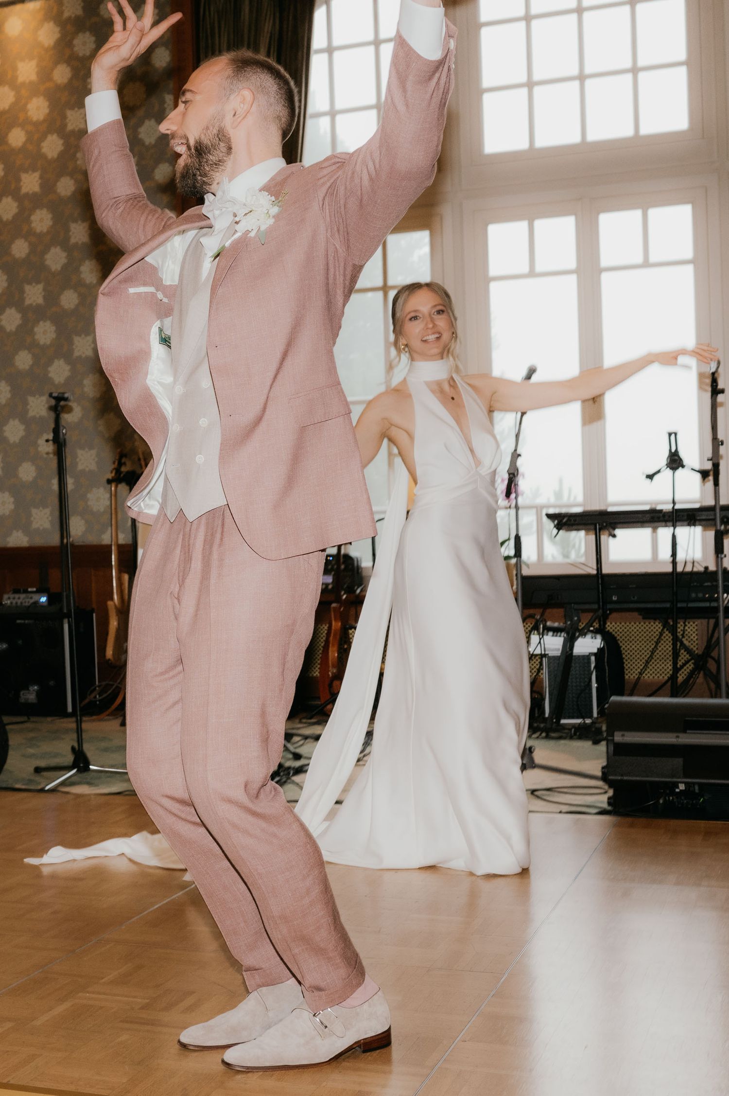 A joyful bride and groom dance energetically at their wedding reception, she in white gown, he in a stylish pink suit.
