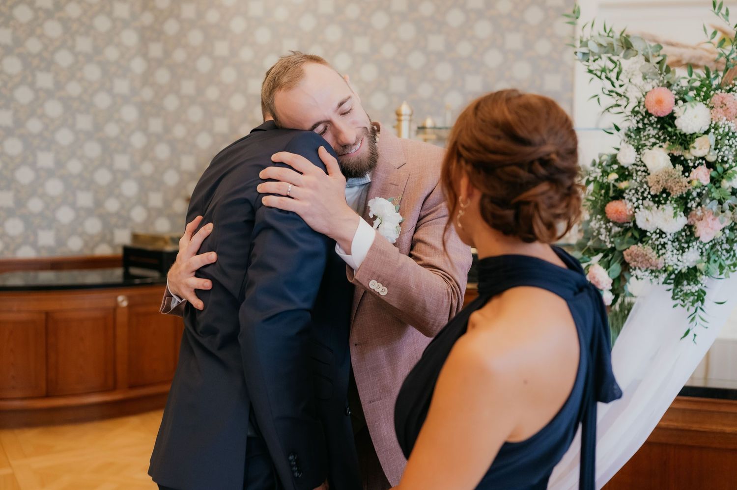 Two men share a warm embrace at a wedding ceremony near a floral arrangement of pink and white roses.