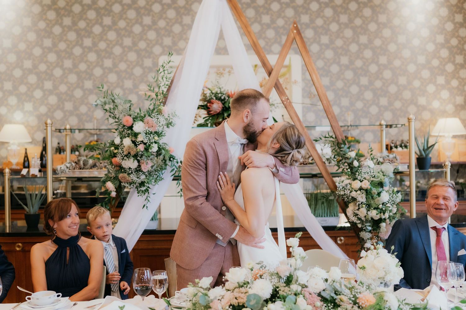 Bride and groom share a kiss at wedding reception table with floral triangle arch backdrop and elegant floral arrangements.