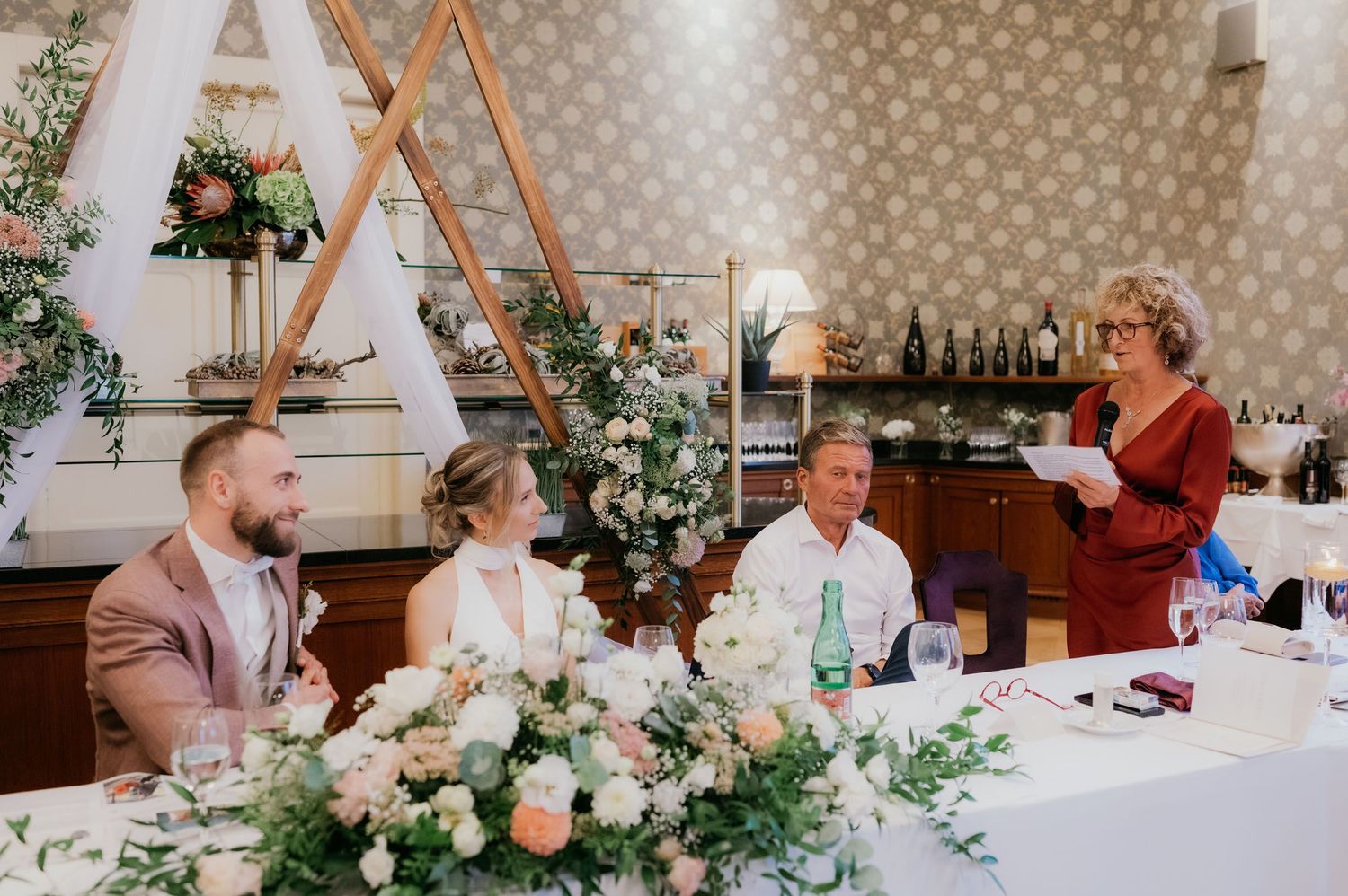 Woman in red dress gives wedding speech at floral-decorated head table with bride and groom seated nearby.