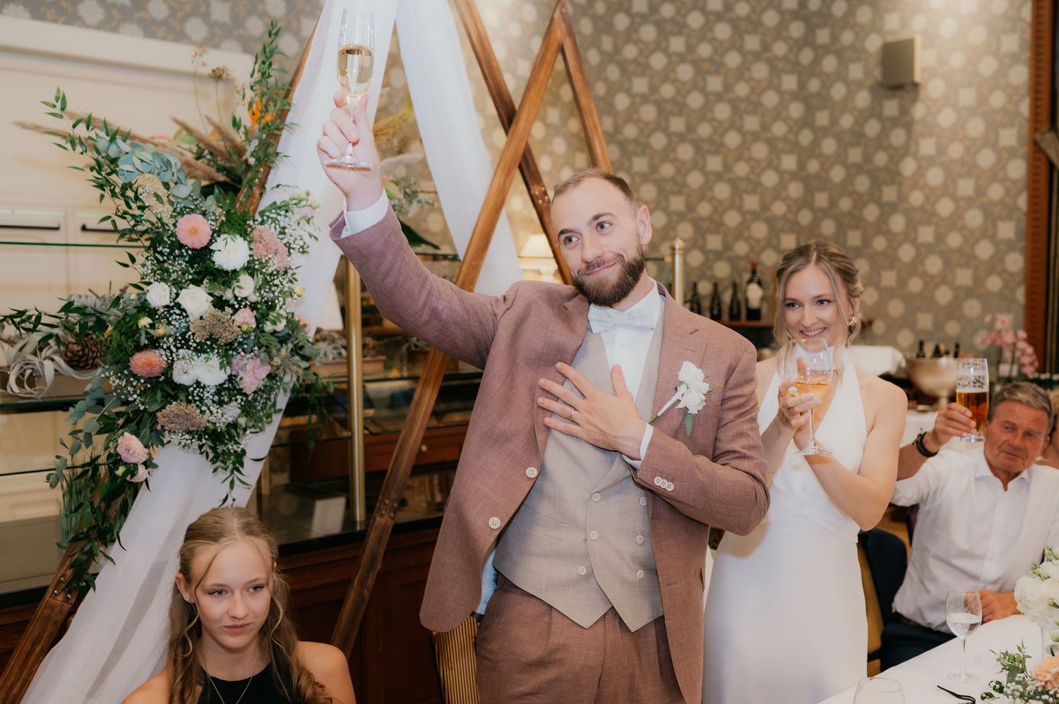 Groom in pink suit raises champagne glass at wedding reception with bride in white dress beside floral arch.