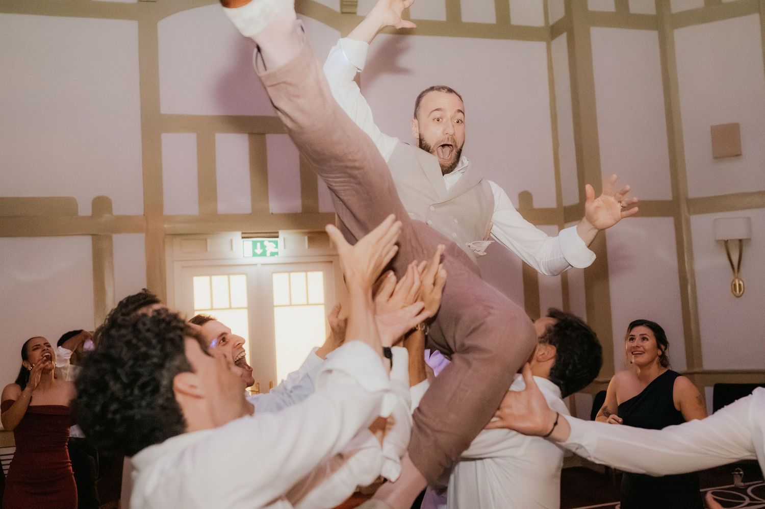 Groom being lifted by wedding guests during energetic reception celebration.