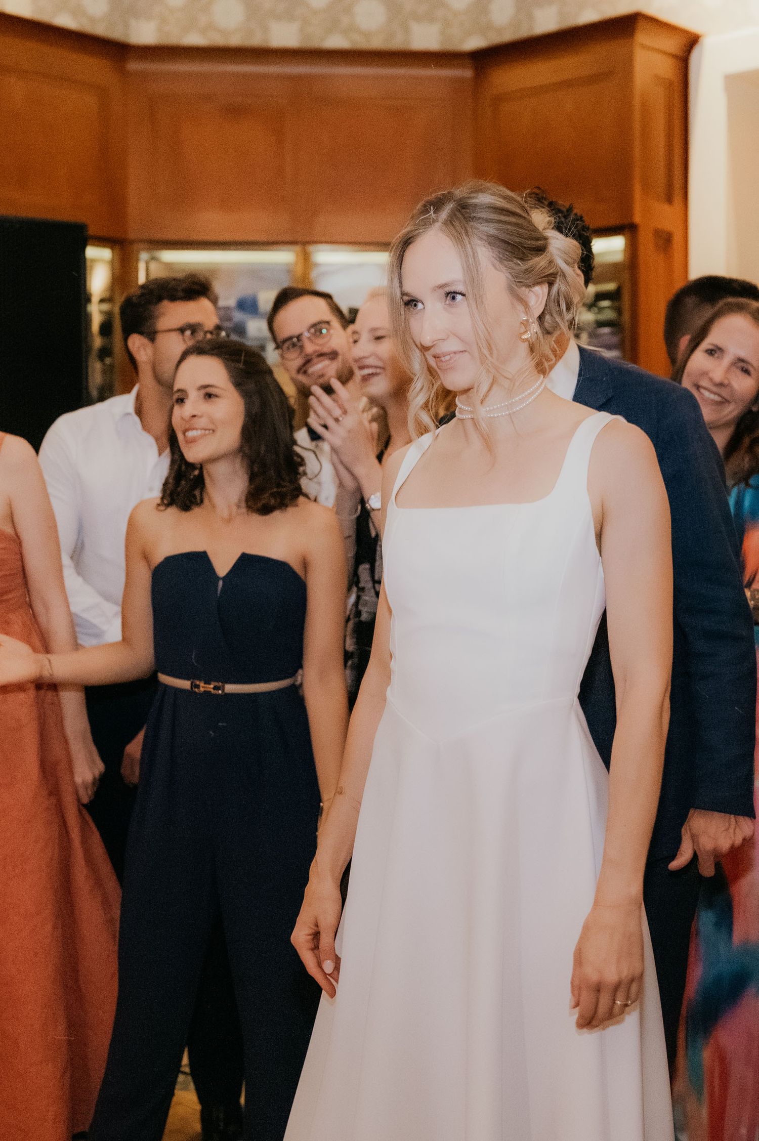 Elegant bride in white gown smiling at wedding reception surrounded by guests clapping in background.