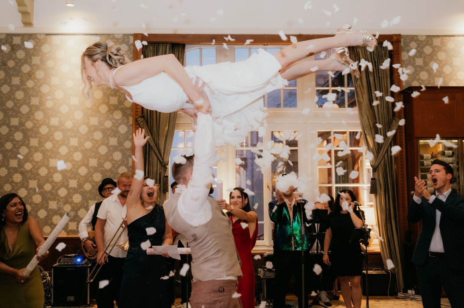 Groom lifts bride during lively wedding reception with confetti and cheering guests in elegant venue.