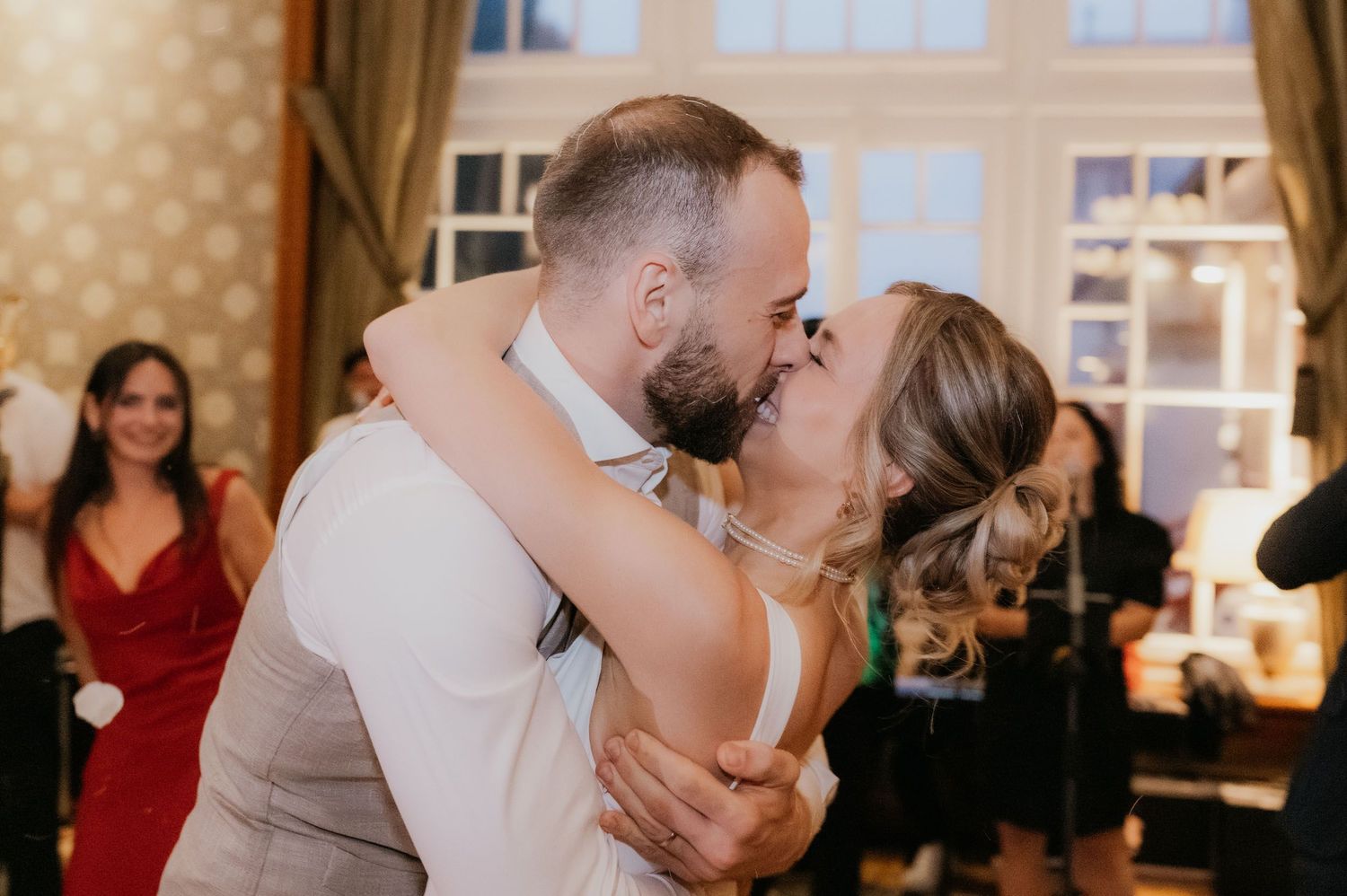 Newlyweds share romantic first kiss during wedding reception while guests celebrate in background.