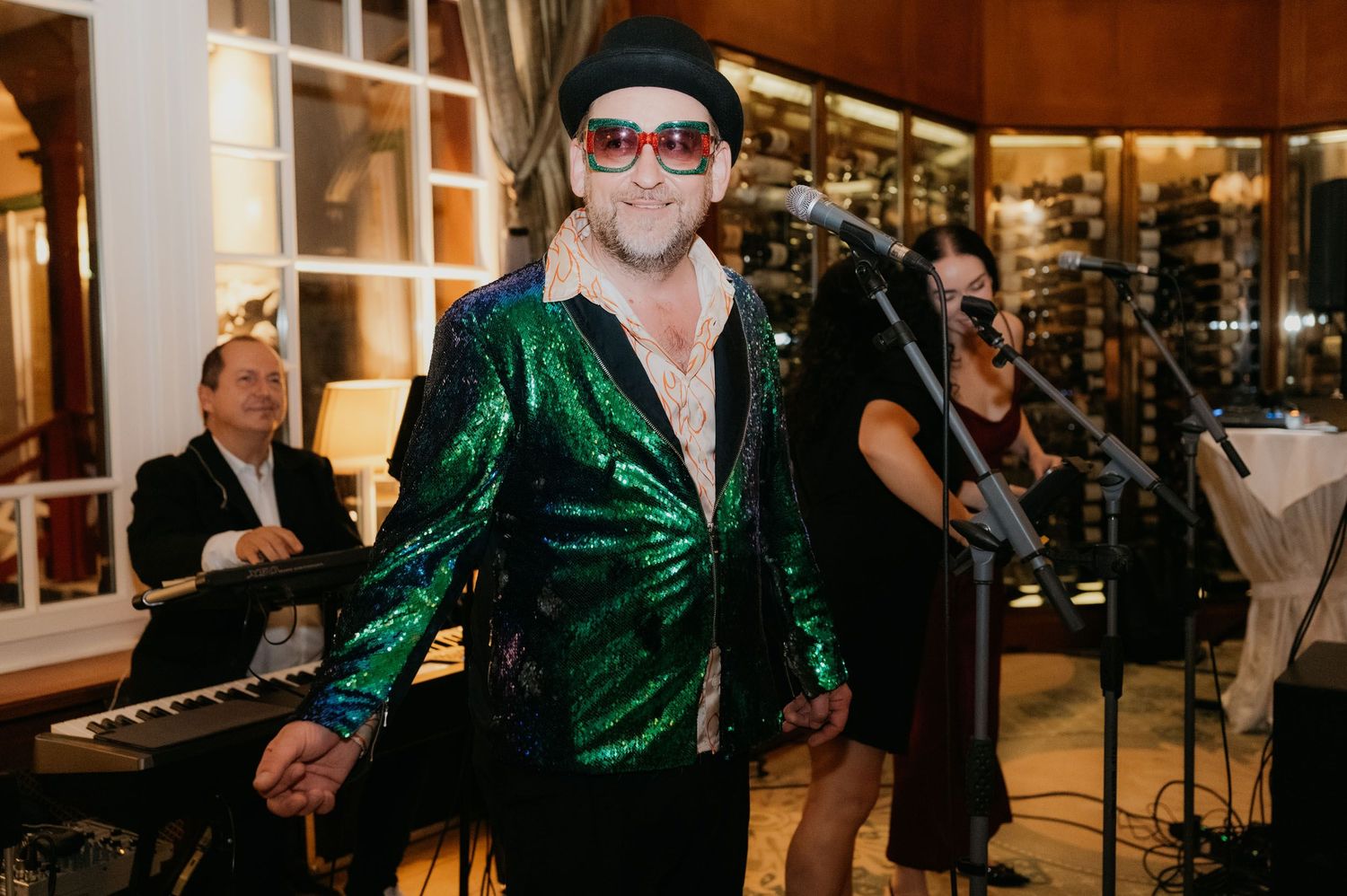 Man in green sequin jacket and sunglasses performs at microphone with band members and wine cellar backdrop.